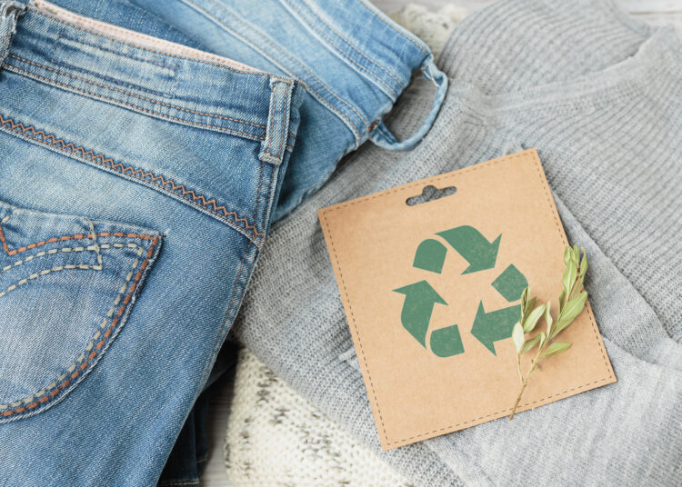 Sustainable still life with Blue denim jeans, gray pullover and craft paper card with Recycling symbol. Second hand apparel idea. Circular fashion, donation, charity concept. Top view